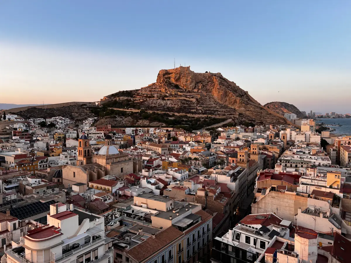 Alicante al atardecer: los tejados del casco antiguo, la cúpula azul de San Nicolás y el castillo de Santa Bárbara iluminado sobre el Benacantil.
